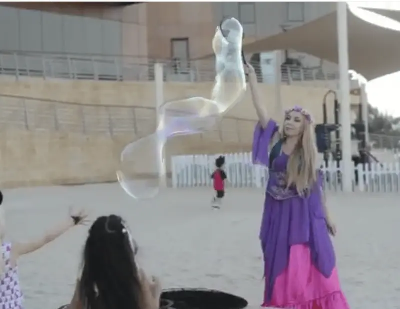 Bubble performer making a long giant bubble on a beach while children reach for it at a family event